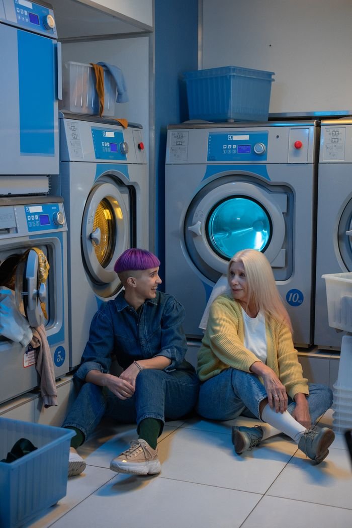 Two women with unique styles sitting on the floor of a laundromat, surrounded by washing machines.