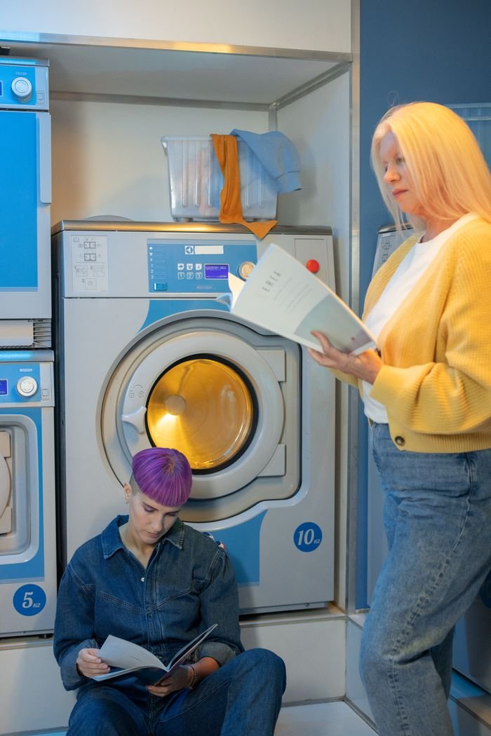 Two adults reading magazines in a busy laundry facility with modern washing machines.