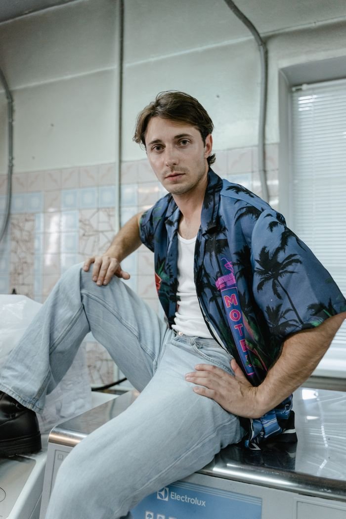 A casually dressed man sits confidently in a laundry room atop a washing machine.